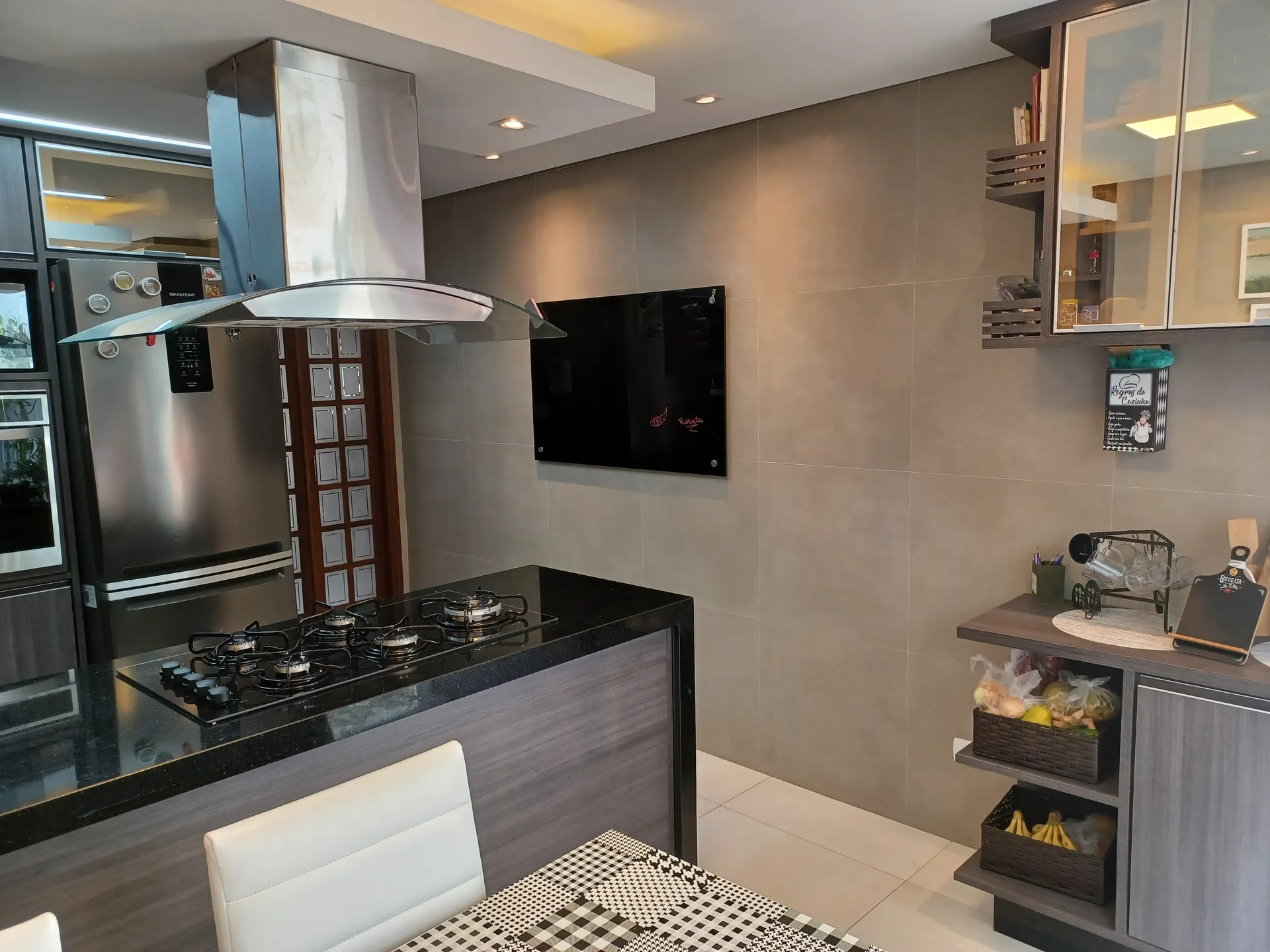 Cozinha de casa com ilha de granito preto, cooktop, coifa inox com vidro e armários aéreos com portas de vidro, projeto residencial da LaArc